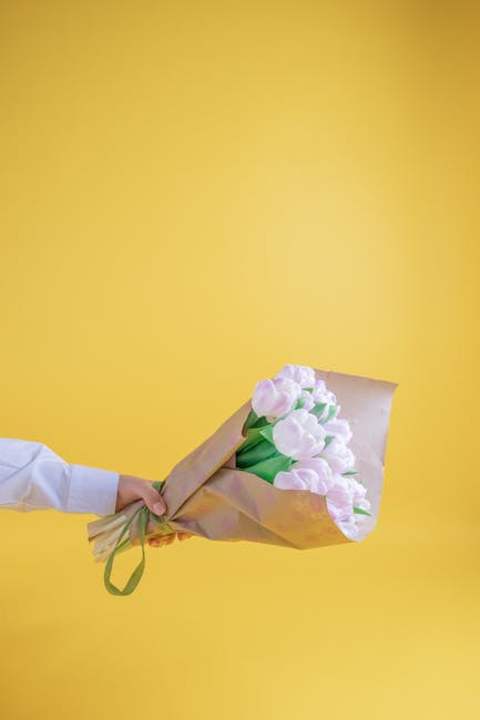 A person wearing a white sleeve is holding a bouquet of light pink tulips wrapped in brown kraft paper, secured with a green ribbon, against a bright yellow background. The tulips have fresh, smooth p