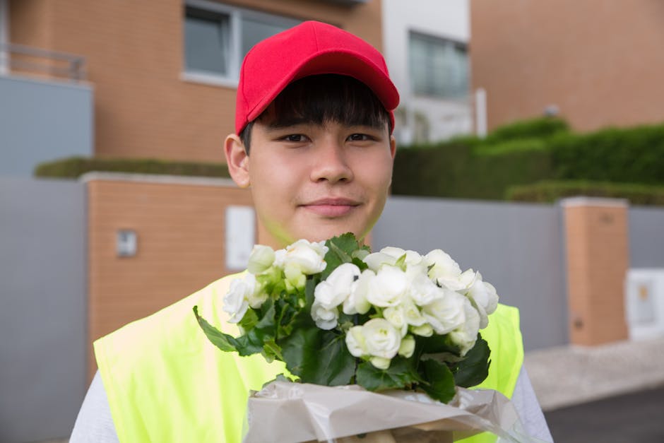 A young delivery person wearing a red cap and a high-visibility yellow vest is holding a floral gift wrapped in white paper with green leaves visible, in front of modern residential buildings with bri