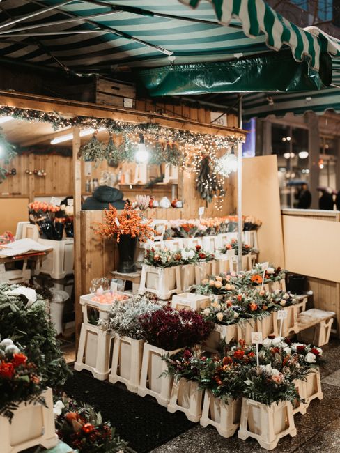 A display of fresh floral arrangements at a market stall, featuring a variety of seasonal flowers in shades of red, pink, white, and orange, carefully arranged in cream-colored containers and small wo