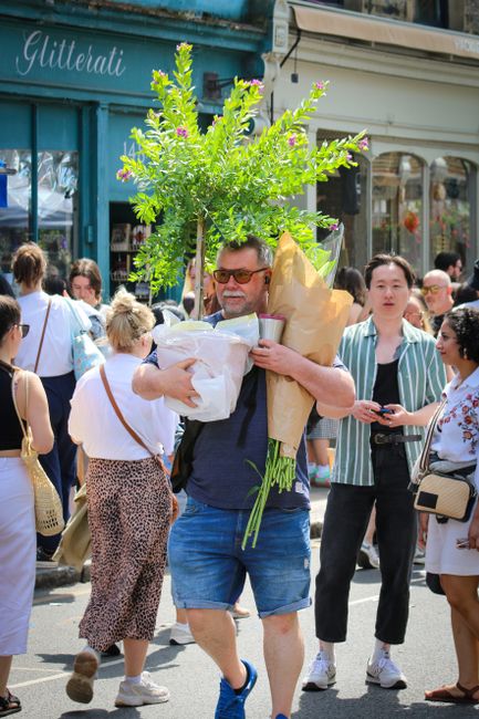 A man holding a large bouquet of fresh flowers, including tall green leafy branches and blossoms with pink accents, wrapped in beige and clear paper, is walking through a busy street scene in Deptford