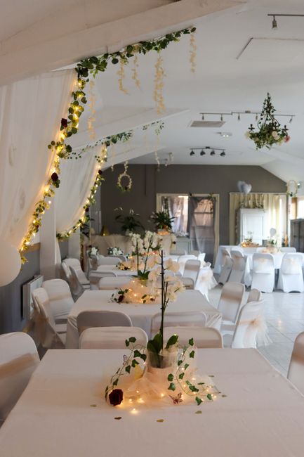 An elegantly decorated wedding reception venue featuring white round tables draped with white tablecloths and surrounded by matching white chairs. Each table has a floral arrangement in a vase, with w